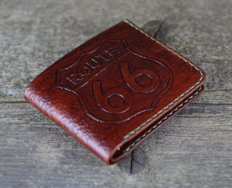 Puede incluir: Una cartera de cuero marr&oacute;n con un borde cosido verde. La cartera presenta un dise&ntilde;o de escudo Route 66 en relieve.