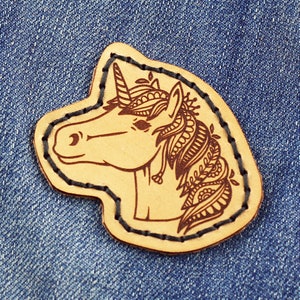 Puede incluir: Un parche de cuero marrón con un contorno negro detallado de un unicornio con un diseño floral. El unicornio tiene un cuerno, una melena y una cola con patrones intrincados.