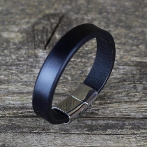 Puede incluir: Una pulsera de cuero negro con un cierre magnético plateado pulido. La pulsera está hecha de una sola banda de cuero negro. El cierre es rectangular y tiene un diseño curvo. La pulsera se muestra sobre una superficie de madera.