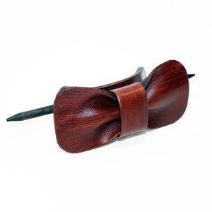 May include: A brown leather hair stick with a bow shape. The stick is made of dark brown leather and has a smooth finish. The bow is tied in a simple knot and has a slightly textured surface.