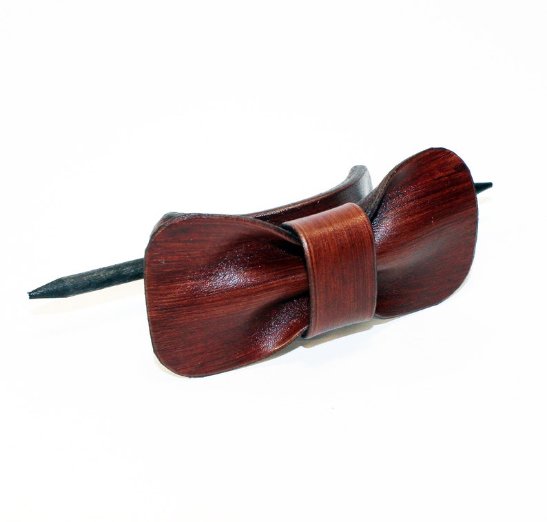 May include: A brown leather hair stick with a bow shape. The stick is made of dark brown leather and has a smooth finish. The bow is tied in a simple knot and has a slightly textured surface.