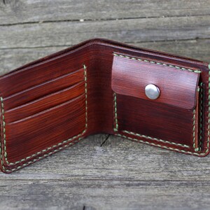 Puede incluir: Una cartera de cuero marr&oacute;n con un borde cosido verde. La cartera tiene m&uacute;ltiples ranuras para tarjetas y un portafolios. La cartera est&aacute; abierta y el interior es visible.