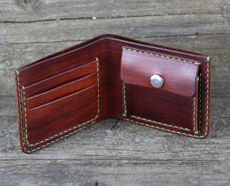 Puede incluir: Una cartera de cuero marr&oacute;n con un borde cosido verde. La cartera tiene m&uacute;ltiples ranuras para tarjetas y un portafolios. La cartera est&aacute; abierta y el interior es visible.