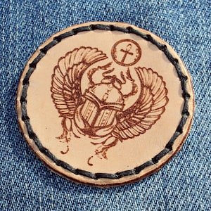 May include: A leather patch with a detailed image of a scarab beetle with wings, surrounded by a black stitched border. The scarab is facing forward with its wings spread out. Above the scarab is a small circle with an ankh symbol inside.