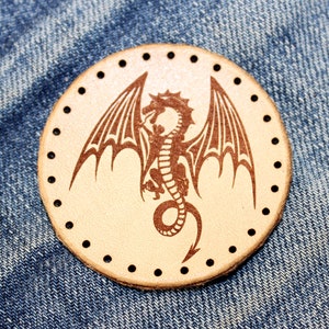 May include: A brown leather patch with a circle of stitching around the edge. The center of the patch features a brown silhouette of a dragon with wings spread wide.