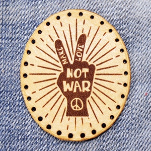 May include: A beige leather patch with a peace sign and the text "Make Love Not War" in brown lettering.