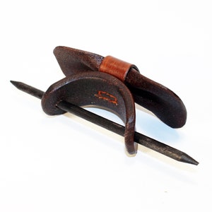 May include: A brown leather hair stick with a brown leather band. The stick is made of metal and has a pointed end. The band is wrapped around the stick and has a decorative stitch.