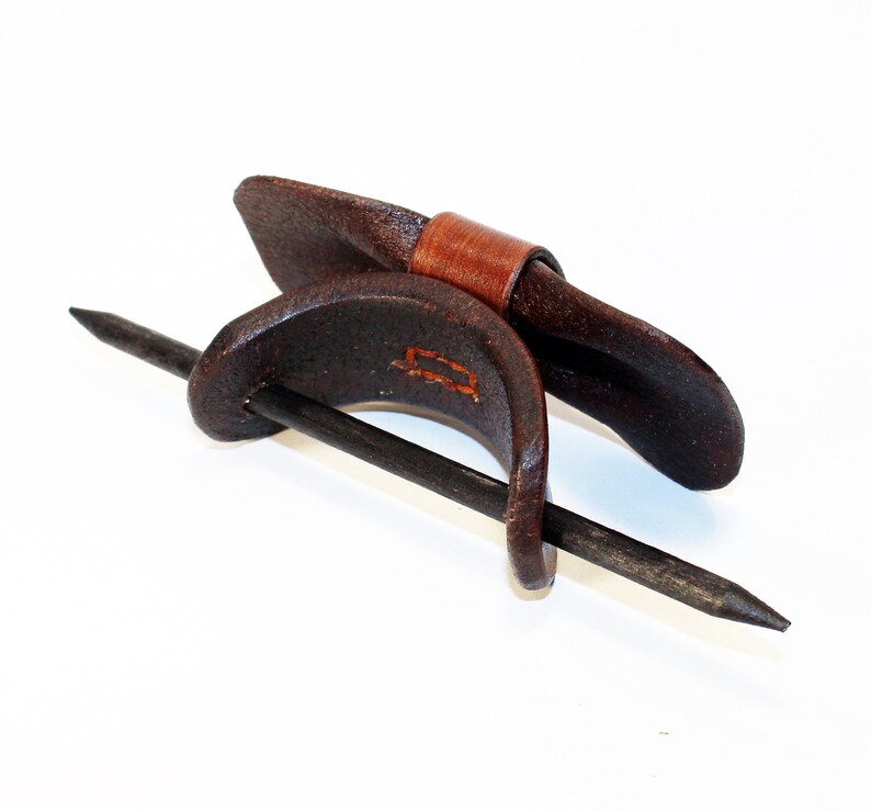 May include: A brown leather hair stick with a brown leather band. The stick is made of metal and has a pointed end. The band is wrapped around the stick and has a decorative stitch.
