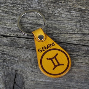 May include: A yellow leather keychain with a silver key ring. The keychain has the Gemini zodiac symbol and the word "GEMINI" engraved on it.