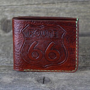 Puede incluir: Una cartera de cuero marr&oacute;n con un dise&ntilde;o de la Ruta 66 en relieve en la parte delantera. La cartera tiene costuras verdes alrededor de los bordes.