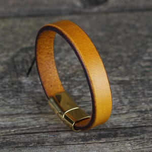 May include: A tan leather bracelet with a gold-coloured magnetic clasp. The bracelet is made of a single strip of leather, with a dark brown edge. The clasp is rectangular and appears to be made of brass.