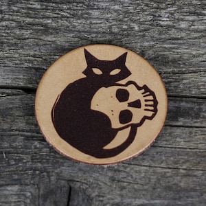 Könnte beinhalten: Ein brauner Lederuntersetzer mit einer schwarzen Silhouette einer Katze, die einen Schädel hält.