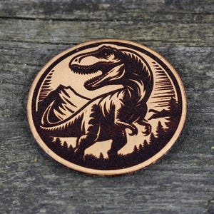 May include: A brown leather coaster with a black silhouette of a dinosaur in a mountain landscape.