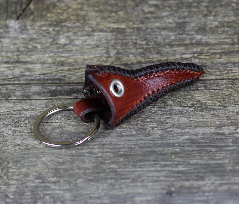 May include: A brown leather key chain with a stitched black border and a metal ring. The leather is shaped like a pointed tip.