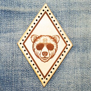 Pode incluir: Um remendo de couro marrom em forma de diamante com um urso marrom usando óculos de sol no centro. O remendo tem costuras ao redor das bordas.