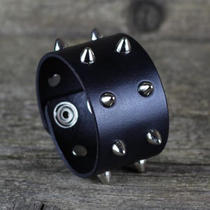 Può includere: Braccialetto in pelle nera con borchie in metallo argentato. Il braccialetto è largo e ha una forma arrotondata.