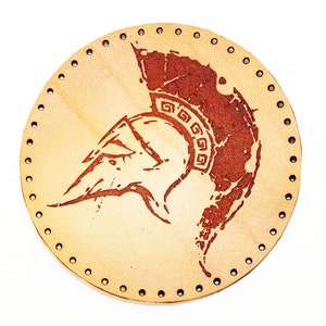 May include: A round, tan leather coaster featuring a brown, distressed design of a Spartan helmet. The helmet has a decorative pattern around the top.