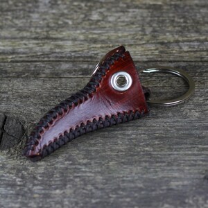 May include: A brown leather keychain shaped like a plague doctor's mask with a silver metal ring. The mask has black stitching around the edges and a silver metal eyelet.