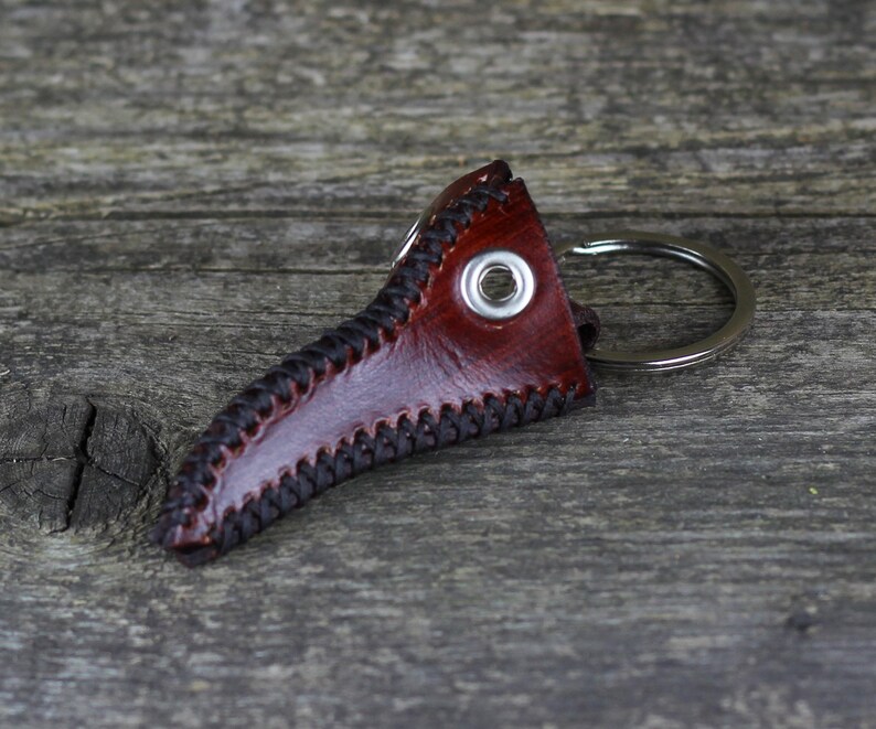 May include: A brown leather keychain shaped like a plague doctor's mask with a silver metal ring. The mask has black stitching around the edges and a silver metal eyelet.