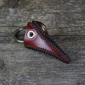 May include: A brown leather plague doctor mask keychain with black stitching and a silver key ring.