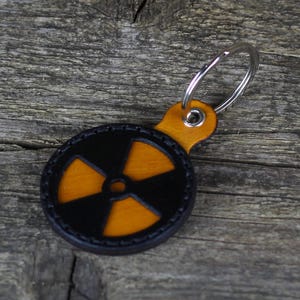 Puede incluir: Un llavero de cuero negro con un símbolo de radiación amarillo. El llavero tiene un anillo de metal plateado.