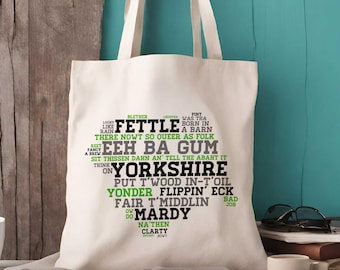 Può includere: Una borsa tote in tela color crema con una grafica a forma di cuore composta da varie parole e frasi del dialetto dello Yorkshire stampate in nero e verde, tra cui "Fettle", "Eeh Ba Gum", "Yorkshire", "Mardy" e "Flippin' Eck".