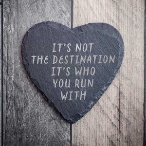 以下が含まれることがあります： 「IT'S NOT THE DESTINATION IT'S WHO YOU RUN WITH」と刻まれた、ハート型のスレートコースター。 コースターは濃いグレーで、グレーと茶色の木目模様の木製表面に置かれています。