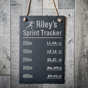 以下が含まれることがあります： ロープハンガー付きの長方形の黒いスレートスプリントトラッカー。「Riley's Sprint Tracker」の文字が上部にあり、距離と時間が下に表示されています。距離は100mから400mまで。時間は分と秒の形式です。