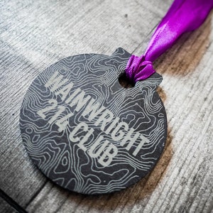 May include: A black round ornament with a topographic map design and the text "Wainwright 214 Club" in white lettering. A purple ribbon is attached to the ornament.