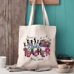 May include: A cream-colored canvas tote bag featuring a colorful graphic print. The design includes silhouettes of people dancing, a boombox, and the text "The best Dance TEACHER Miss Smith" in a multi-colored leopard print and gradient font.