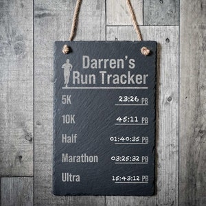 Könnte beinhalten: Eine rechteckige Schiefertafel in Schwarz mit einer Aufhängung aus Juteseil. Die Oberfläche zeigt den eingravierten Text 'Darren's Run Tracker', ein Läufer-Symbol und eine Liste von Laufdistanzen wie 5K, 10K, Half, Marathon und Ultra mit Platzhaltern für persönliche Bestzeiten.