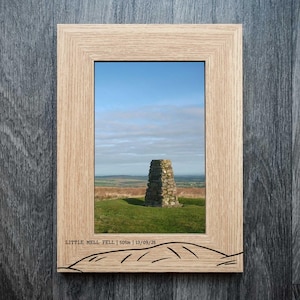 May include: A framed photograph of Little Mell Fell, showing a stone cairn on a grassy hill under a blue sky. The frame is made of light-coloured wood. The text "LITTLE MELL FELL | 505m | 13/09/25" is printed on the frame.