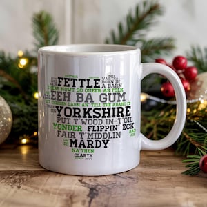 Könnte beinhalten: Weißer Keramikbecher mit schwarzem und grünem Text, darunter das Wort "Yorkshire" und regionale Ausdrücke. Der Becher steht auf einer Holzoberfläche, mit Weihnachtsdekoration im Hintergrund.
