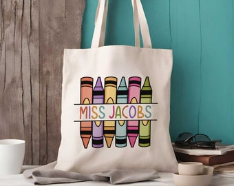 May include: A cream-colored canvas tote bag featuring a graphic print of six colorful crayons arranged in a row. The text MISS JACOBS is printed in a multi-colored sans-serif font across the center of the crayon illustration.