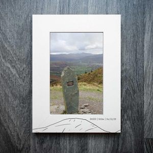May include: A framed photograph of a mountain vista. The image shows a stone marker with a plaque, set against a backdrop of hills and a cloudy sky. The white frame has a mountain outline. Text reads: DODD | 502m | 14/12/25.