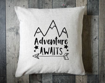 May include: White throw pillow with black text that reads "Adventure Awaits" with a mountain range graphic and an arrow pointing to the right.