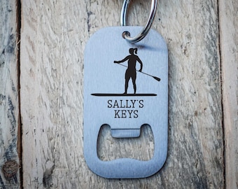 以下が含まれることがあります： パドルボードをしている人のシルエットと「SALLY'S KEYS」の文字が刻まれたシルバーのボトルオープナーキーホルダー。