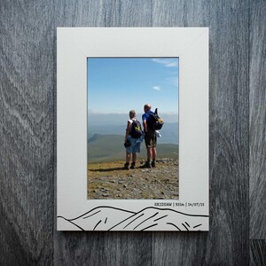 May include: White framed photograph showing two people on a mountain summit, looking out at a vista under a blue sky. The frame has a black mountain outline and the text "SKIDDAW | 931m | 14/07/15".