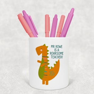 May include: White ceramic pencil holder with a cartoon dinosaur illustration. The dinosaur is orange and green with a white background. The text on the holder reads "Mr. Rowe is a roarsome teacher!"