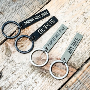 May include: Keychains with rectangular metal tags and circular key rings. One black keychain displays "CARDIFF HALF 2025", another shows "01:34:15", and a third reads "ANY RACE". The keychains are on a wooden surface.