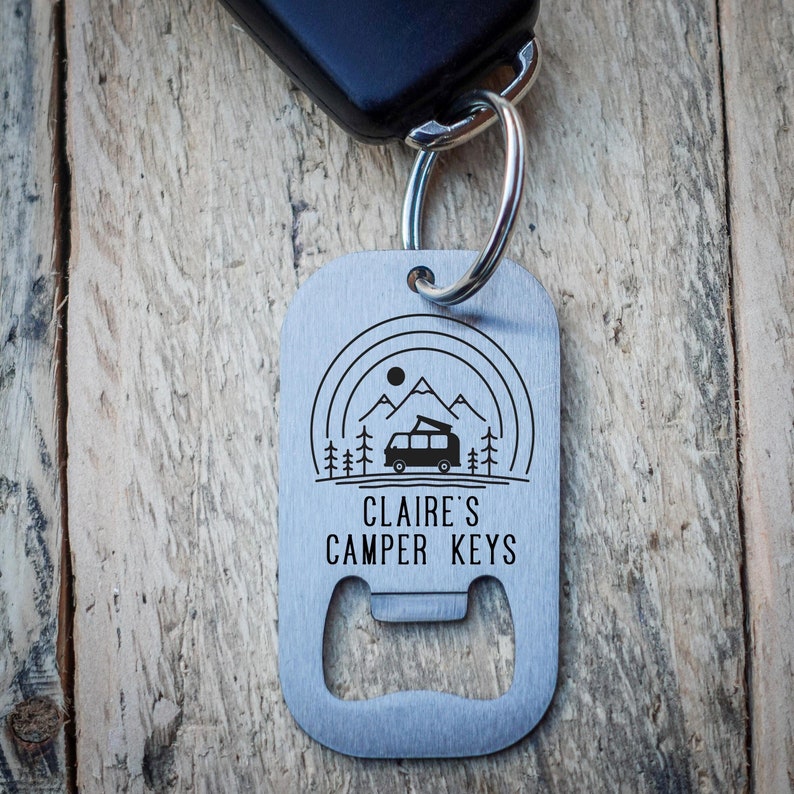 Puede incluir: Un llavero de metal plateado con un abridor de botellas. El llavero presenta un gr&aacute;fico de una autocaravana frente a monta&ntilde;as con el texto "CLAIRE'S CAMPER KEYS".