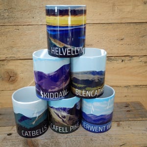 May include: A stack of six white ceramic mugs with landscape designs. Each mug features a different mountain scene with names like "Helvellyn" and "Skiddaw" printed on them. The mugs have a glossy finish and are stacked on a wooden surface.