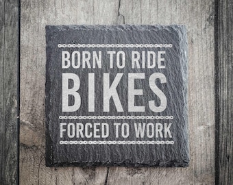 May include: A square, dark grey slate coaster with the text "BORN TO RIDE BIKES FORCED TO WORK" in white, framed by a bicycle chain design. The coaster is on a weathered wooden surface.
