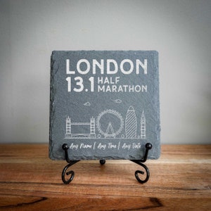 Könnte beinhalten: Eine graue Schiefertafel mit einer weißen Umrandung der Londoner Skyline, einschließlich Big Ben, dem London Eye und anderen Sehenswürdigkeiten. Der Text "LONDON 13.1 HALF MARATHON" ist in Weiß auf die Tafel gedruckt. Die Tafel steht auf einem schwarzen Metallständer. "Any Name | Any Time | Any Date" ist unter der Skyline gedruckt.