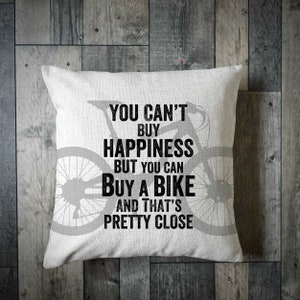 May include: Square white throw pillow with a bicycle silhouette and the text "YOU CAN'T BUY HAPPINESS BUT YOU CAN BUY A BIKE AND THAT'S PRETTY CLOSE" in black. The pillow is on a wooden surface.