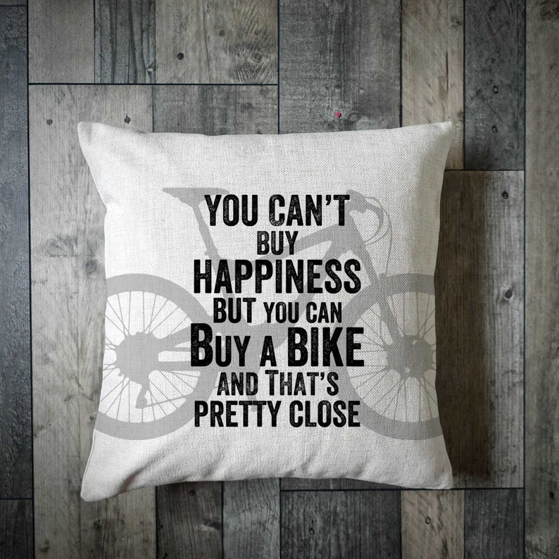 May include: Square white throw pillow with a bicycle silhouette and the text "YOU CAN'T BUY HAPPINESS BUT YOU CAN BUY A BIKE AND THAT'S PRETTY CLOSE" in black. The pillow is on a wooden surface.