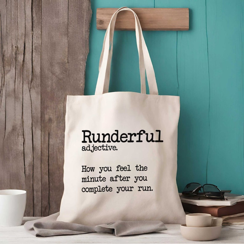 Puede incluir: Una bolsa de tela de color crema con dos correas para el hombro. La parte frontal presenta un texto impreso en negro que dice: Runderful adjective. How you feel the minute after you complete your run. La bolsa tiene una forma rectangular.