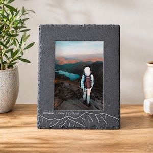 May include: A dark grey slate photo frame displaying a landscape image of a hiker on a mountain. The frame is inscribed with "SNOWDON | 1085m | 13/06/26" and a mountain range design. The frame is made of slate.