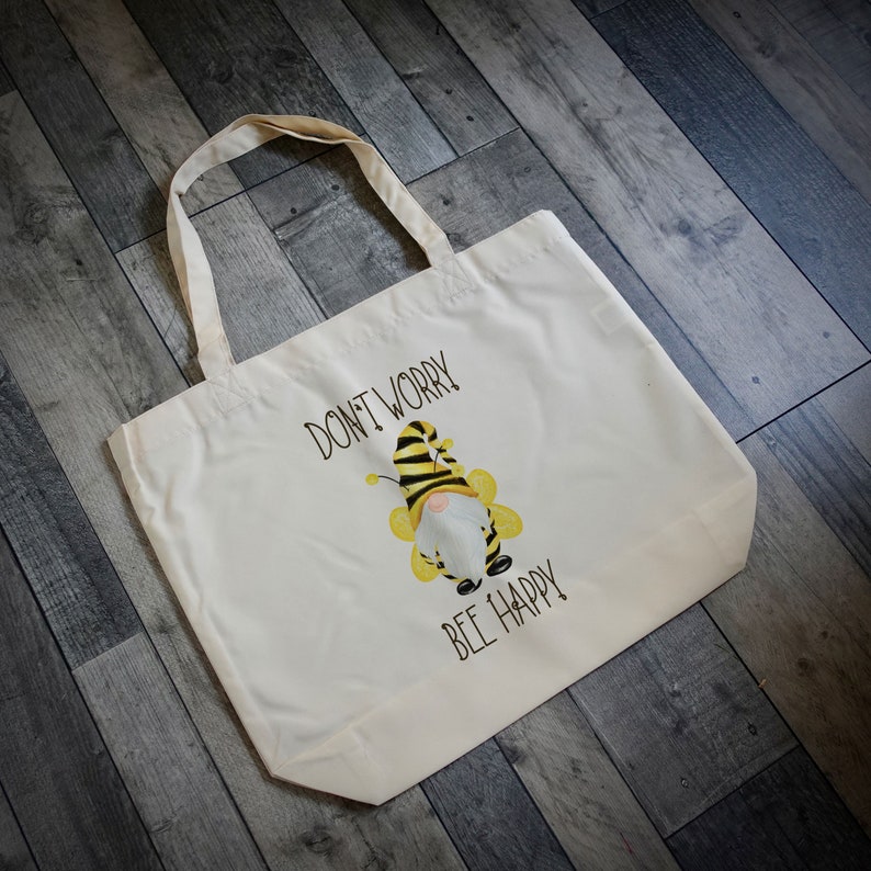 May include: Cream-colored tote bag with black text that reads "DON'T WORRY BEE HAPPY." The bag features a whimsical illustration of a gnome dressed as a bee, with yellow and black stripes. The bag has two handles.
