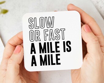 May include: A square white coaster with rounded corners featuring black text that reads SLOW OR FAST A MILE IS A MILE. The top two lines are in a thin outline font, while the bottom two lines are in a bold, solid black sans-serif typeface.